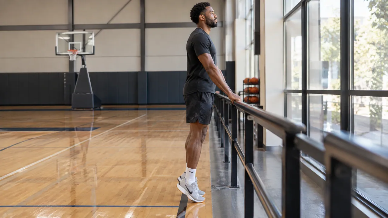 Basketball player using a court-side rail for balance during a controlled calf raise